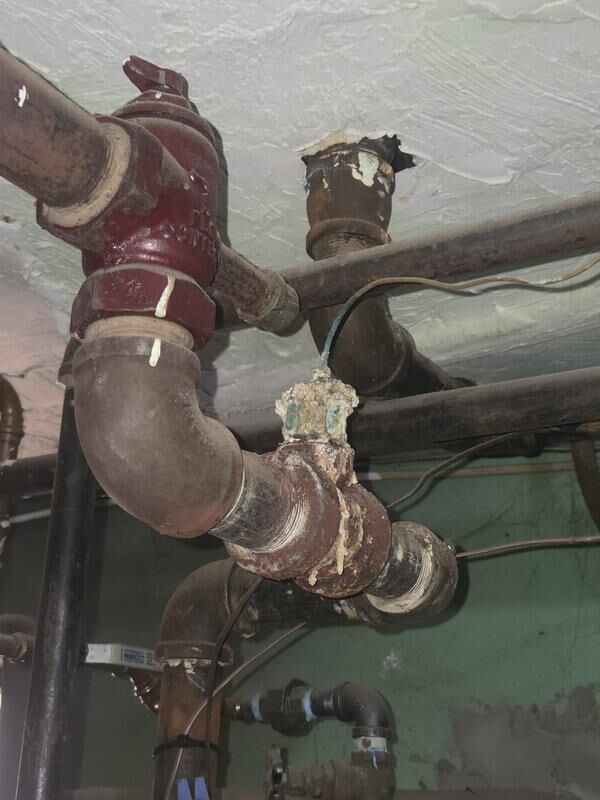 Conducted inspection of commercial boiler system showing extensive deterioration of pipe infrastructure. Documented severe corrosion at multiple connection points, including a compromised reddish valve assembly and significant mineral buildup at joints. Noted structural damage to wall penetration with visible cracking extending from pipe entry point, requiring both immediate repair options and complete system replacement alternatives for client consideration.
